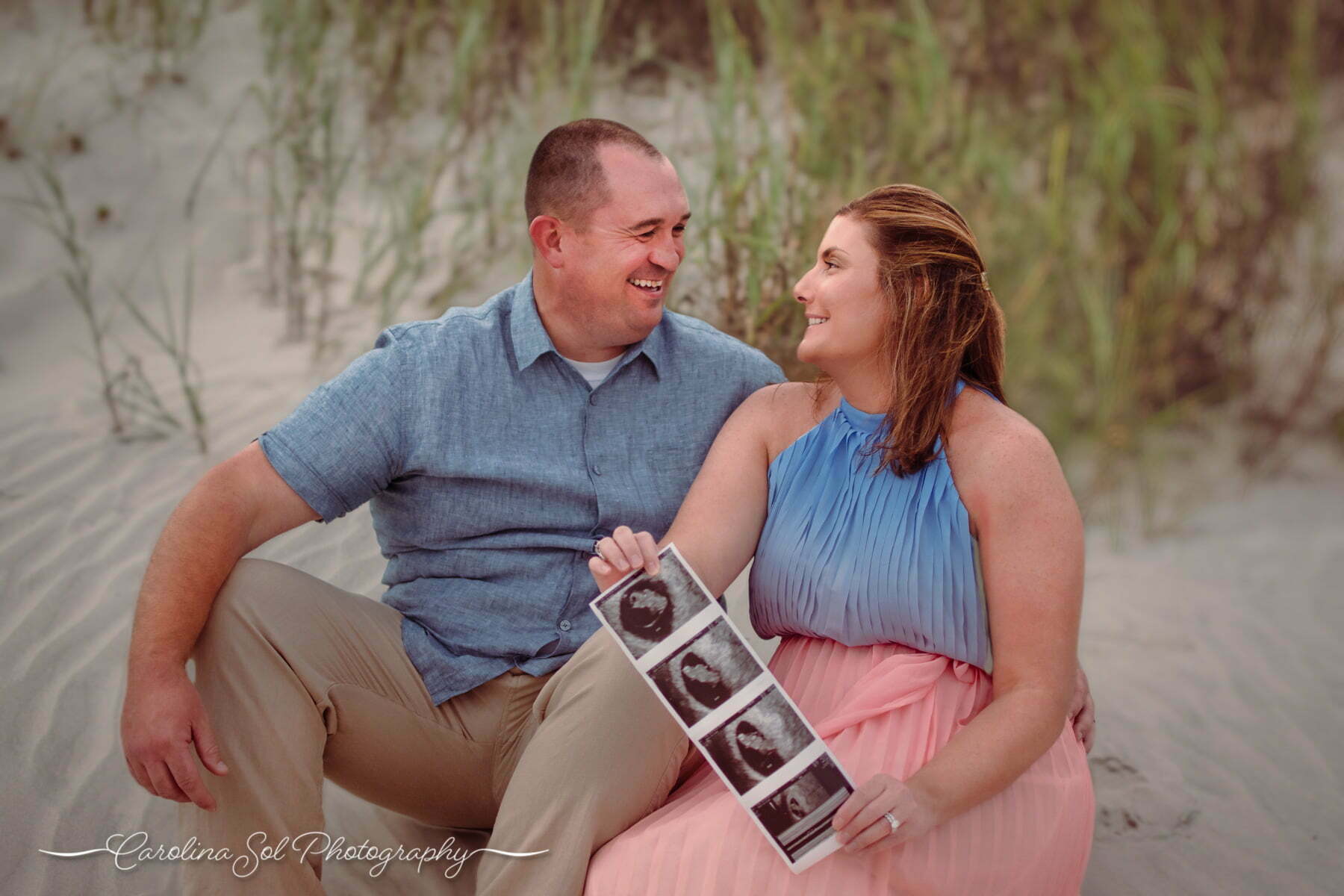 Excited couple relaxing near sand dunes holding ultrasound pictures for pregnancy announcement photoshoot at Sunset Beach, NC.