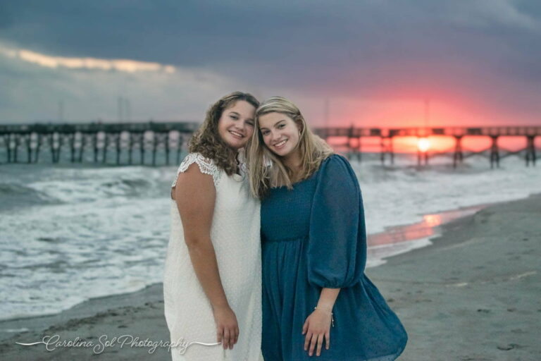 Sisters on vacation at Oak Island Pier smiling for the photographer against a colorful sunset.